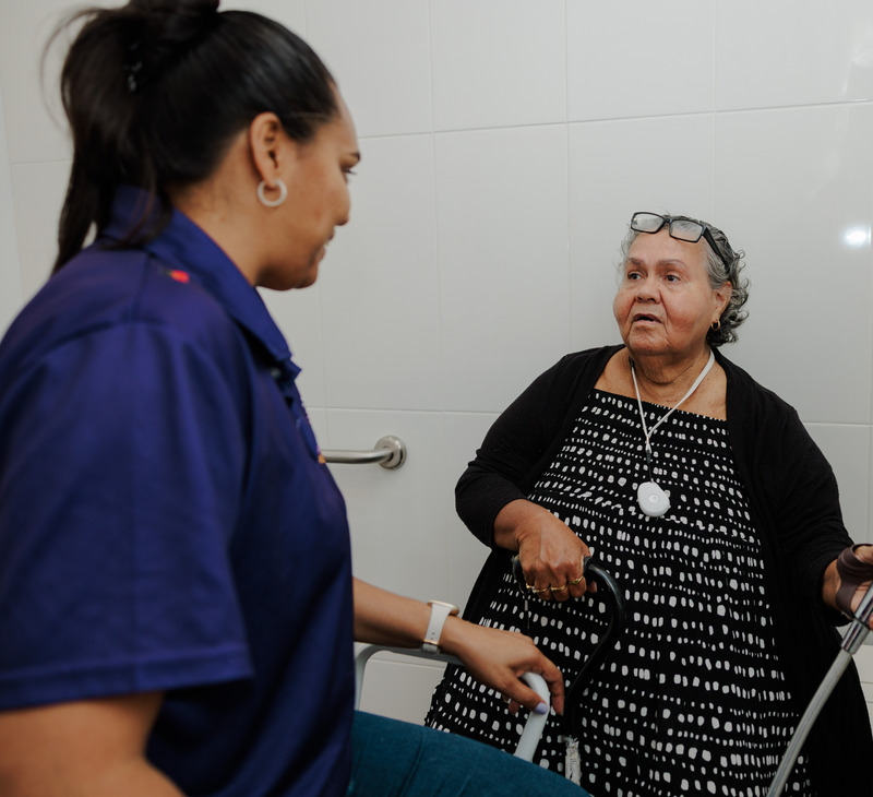 A caregiver assists an older woman with gray hair and glasses, who is holding a cane, in a bathroom with white tiled walls and a grab bar—demonstrating valuable occupational therapy for mobility support.