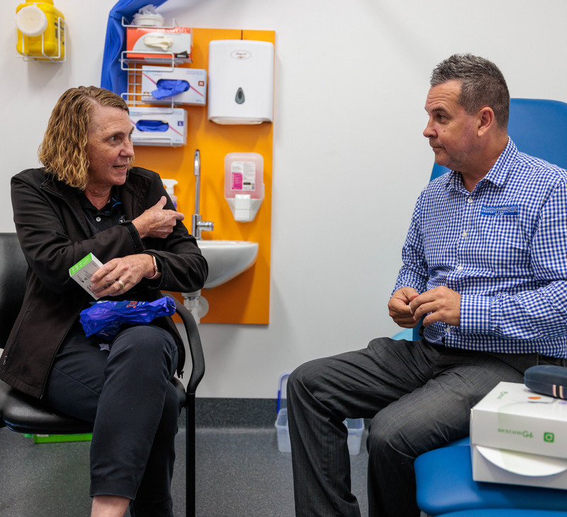 Two people sit in a medical office. One points to their upper arm, possibly discussing a medical issue or vaccination related to diabetes education, while the other listens attentively. Medical supplies are visible on the wall behind them.
