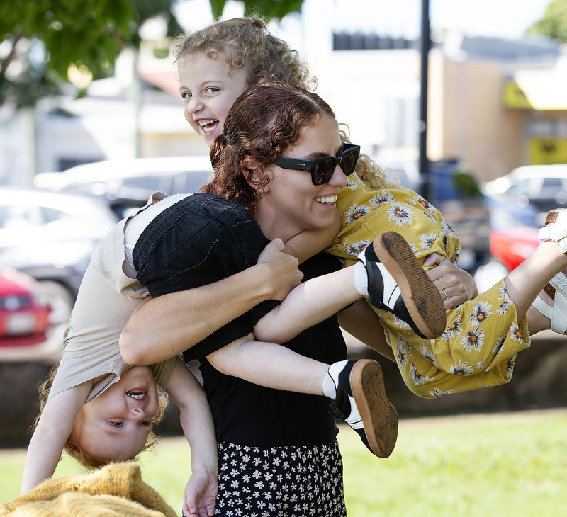 A woman wearing sunglasses smiles while holding two laughing children, one on each shoulder, outdoors in a park. The children are playfully hanging with their legs in the air, capturing a joyful moment that reflects the supportive spirit of Indigenous family wellbeing services.