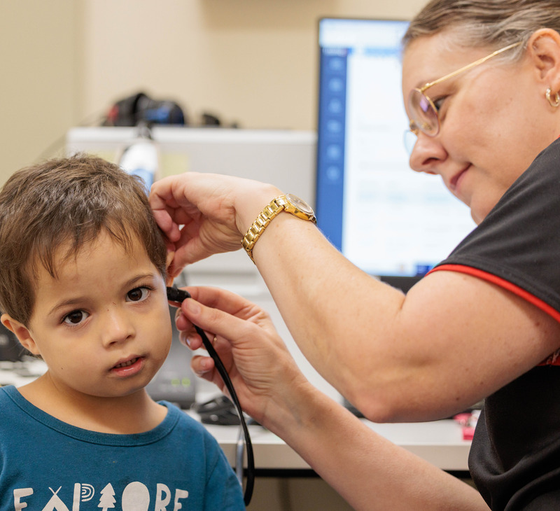 A healthcare worker examines a young boy’s ear with an otoscope in a medical office, promoting ear health for Mob. The boy looks at the camera while the professional focuses on the procedure, and a computer screen is visible in the background.