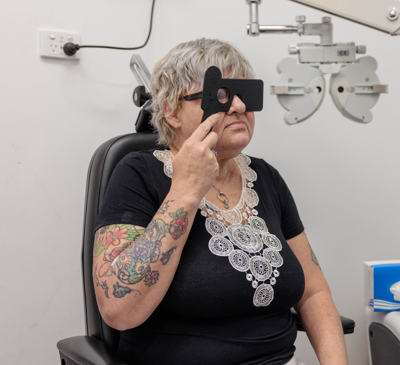 An older woman sits in an eye exam chair, holding a vision testing occluder over her eyes. She has short gray hair, glasses, and floral tattoos on her arms. Ophthalmology equipment is visible in the background, highlighting the importance of eye health for Mob during routine eye checkups.