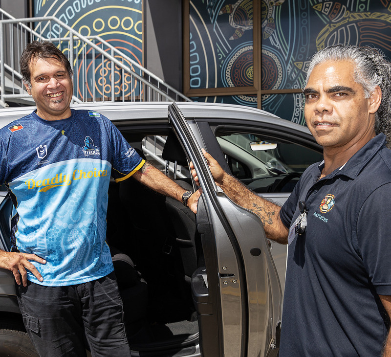 Two men stand beside an open car door, smiling. One leans on the car, wearing a blue sports shirt; the other, in a dark polo shirt, holds the door—perhaps ready to offer friendly transport services for Mob. Colorful artwork is visible on the wall behind them.