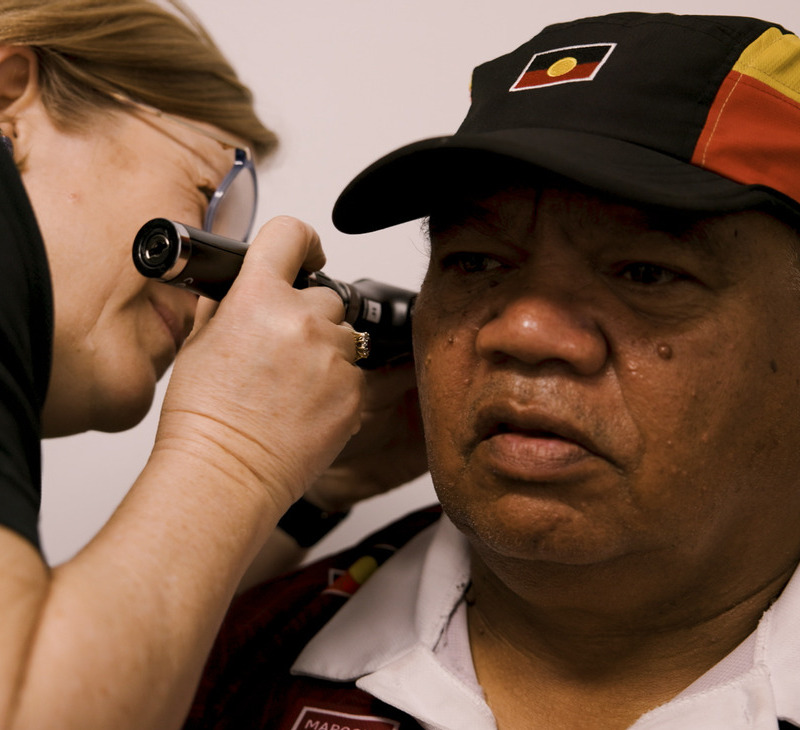 A healthcare worker examines the ear of an Indigenous man wearing a cap with an Aboriginal flag design, as part of a 715 Health Check.
