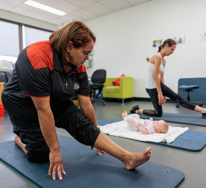 Two women stretch on yoga mats in a bright room as part of a Deadly Fit Mums session. One woman is near a baby lying on a blanket. There are chairs, a green couch, and wall decorations in the background.