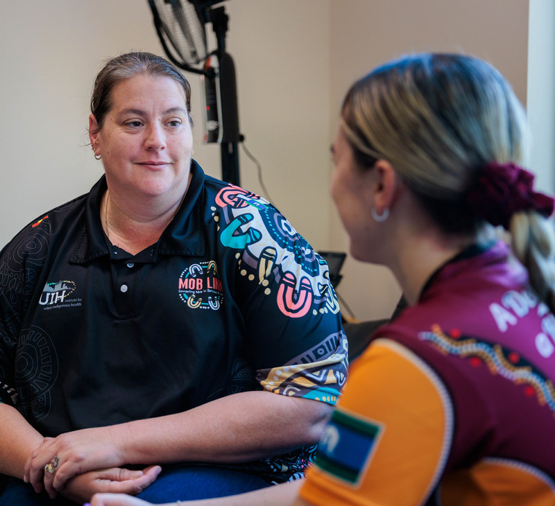 Two women sit facing each other in conversation, reflecting on aspects of care coordination. One wears a black shirt with colorful patterns and logos, while the other has a maroon shirt with similar patterns. They appear to be in a casual indoor setting.