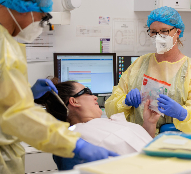 Two healthcare workers in PPE assist a patient in a dental chair, ensuring the highest standards of dental and oral health for Mob. One holds dental tools in a bag, while the other stands nearby. A computer with a chart is visible in the background.