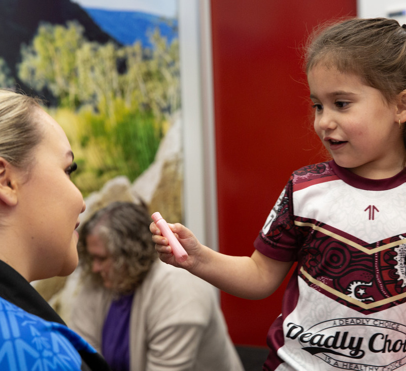 A young girl in a maroon and white shirt holds a pink marker and smiles, interacting with a seated woman. The joyful scene reflects the inclusive spirit fostered by disability services for Mob. A blurred person sits in the background with green and red walls behind them.