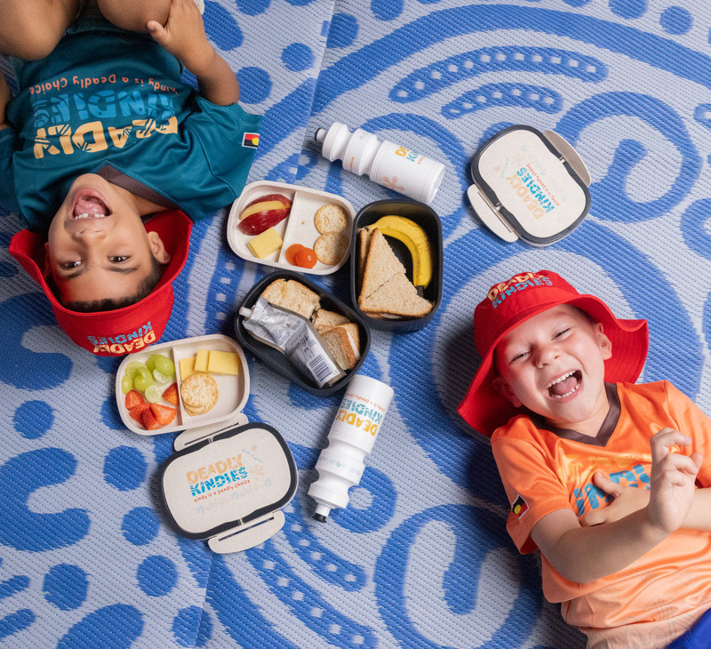 Two smiling children lie on a blue patterned mat with lunchboxes of healthy snacks, sandwiches, fruit, and water. Both wear red hats and colorful shirts, enjoying a playful moment together that reflects the fun and nourishment central to Early Childhood Education.