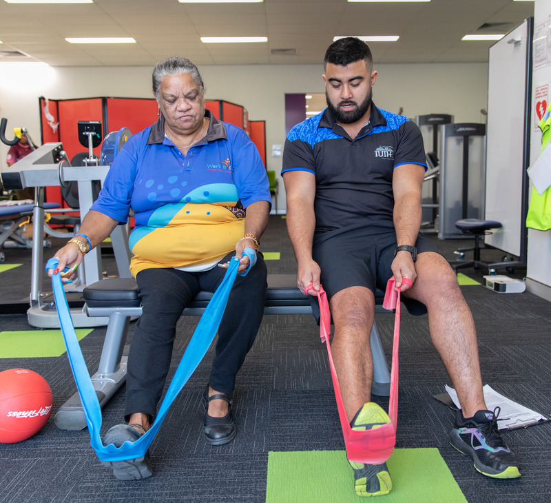 An older woman and a man sit on a bench using resistance bands for foot exercises in a gym, focusing on rehabilitation or physical therapy activities rooted in exercise physiology for mobility improvement.