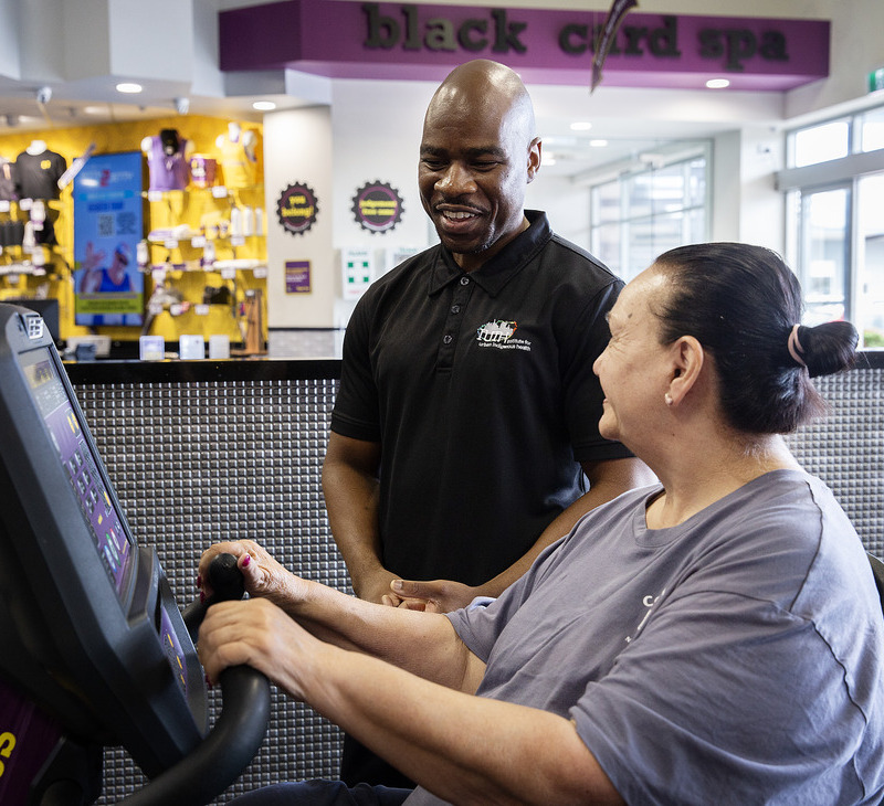 A woman uses a cardio machine at a gym while a staff member in a black polo shirt stands nearby, smiling and offering support. In the background, "Black card spa" is visible, creating an inviting atmosphere ideal for heart rehab for Mob.