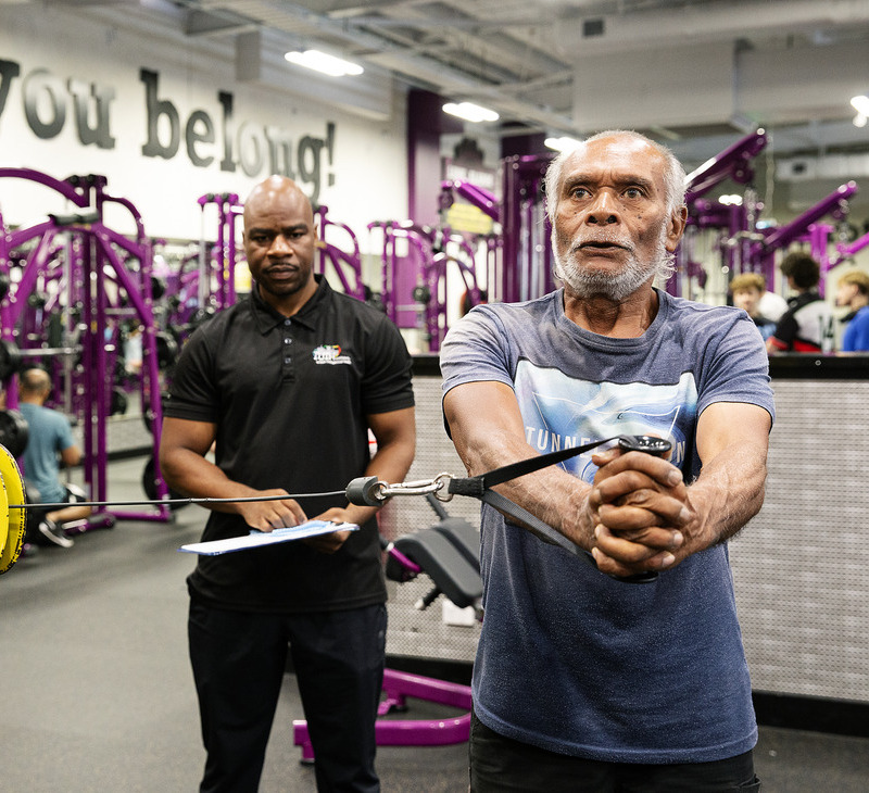 An older man uses a cable machine for exercise in a gym while a trainer watches and takes notes, focusing on lung rehab for Mob. The gym has purple equipment and a sign on the wall reads, "You belong!.