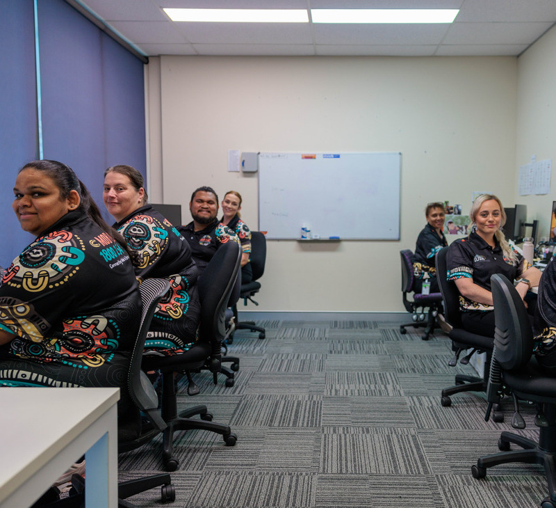 Seven people sit at desks in an office, smiling at the camera. They wear matching black shirts with colorful patterns featuring the Mob Link logo. The room has blue blinds, a whiteboard, and gray carpet.