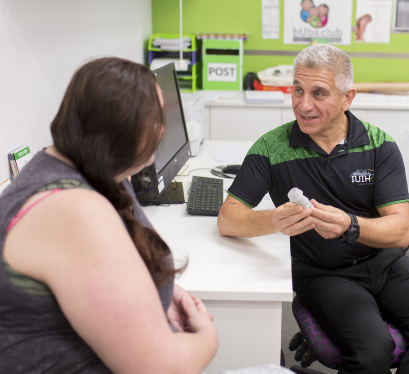 A healthcare professional talks to a seated woman in an office, holding a medical device while discussing persistent pain services for Mob. Both appear engaged in conversation. A computer and office supplies are visible in the background.
