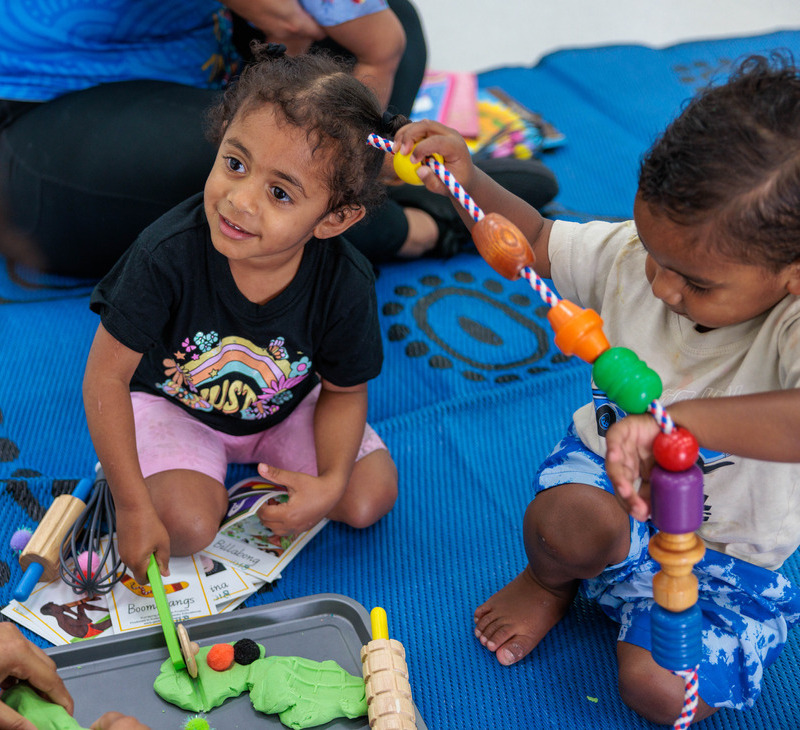Two young children sit on a blue mat at MATSICHS Playgroups, playing with green playdough and colorful toys. One child smiles up while holding a tool, and the other concentrates on a beaded stick.