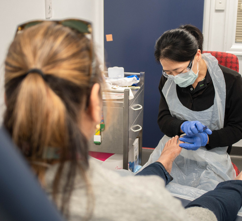A healthcare worker wearing gloves and a mask examines a seated patient's bare foot in a medical clinic, focusing on foot health for mob. Medical supplies and equipment are visible in the background.