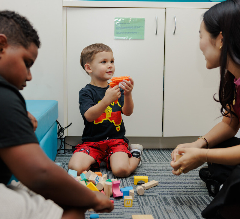 Three children sit on the floor indoors, playing with colorful wooden blocks. As they build and explore, one child holds up a block and looks at an adult nearby—who offers speech and language help for Mob while smiling and engaging in the activity.