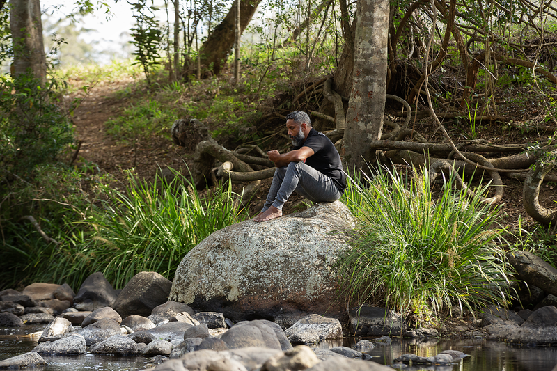 Image of Staying Deadly mental health ambassador, Preston Campbell, sitting on rocks with a flowing creek running nearby.