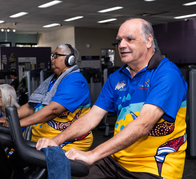 Two older adults in colorful shirts exercise on stationary bikes at a gym; one wears headphones and a towel around her neck, while the other smiles, both focused on their workout as part of an exercise rehab for mobility program.