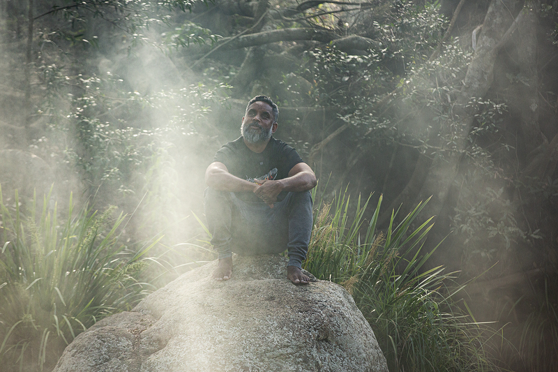 Preston Campbell sits on a rock in a rainforest like setting, the light is shining down through the trees and surrounding him on the rock.