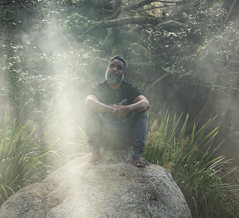 A person with a beard sits barefoot on a large rock, surrounded by mist and lush greenery in a forest. Sunlight filters through the trees, creating a peaceful, serene atmosphere—a quiet moment reflecting on staying deadly and nurturing mental health amidst nature’s calm.