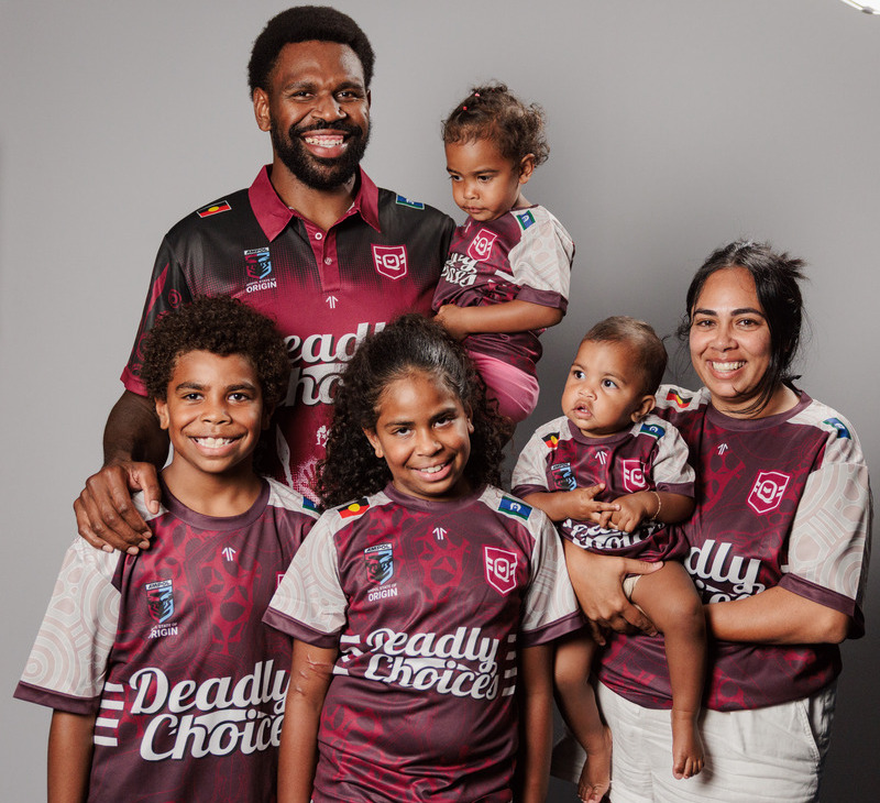 A smiling family of six poses together in matching maroon and white sports jerseys reading "Deadly Choice" against a plain background. Two adults and four children stand close, with one child held in each adult's arms, showcasing the spirit of Deadly Dads as they celebrate unity and pride.