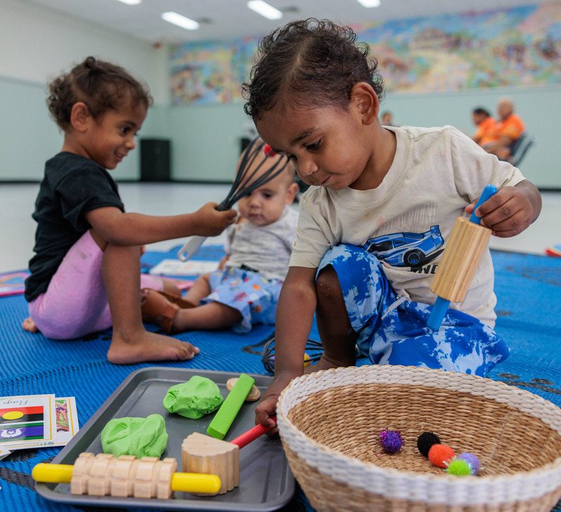 Three jarjums play with wooden toys and craft materials on a blue mat indoors in a warm, supportive space inspired by the values of the Birthing in Our Community (BiOC) program.