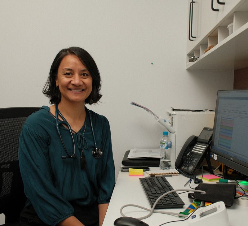 A doctor with a stethoscope around her neck sits smiling at a desk in a medical office, representing the dedicated BiOC GP services at Moreton ATSICHS, with a computer, phone, and office supplies visible around her.