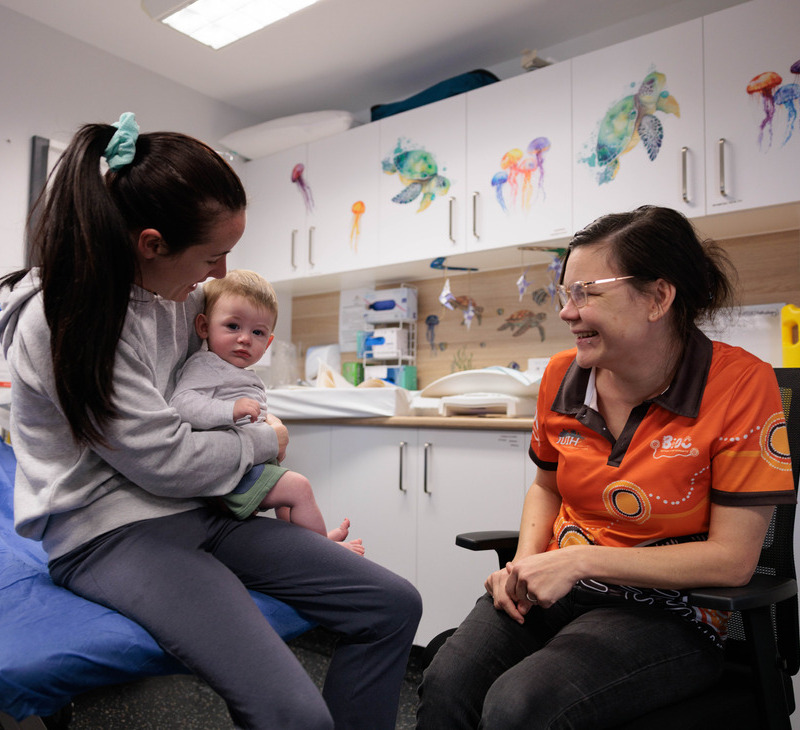 A woman holds a baby on her lap while smiling and talking to a BiOC Child Health worker in an orange shirt, inside a colorful medical clinic room decorated with sea animal illustrations.
