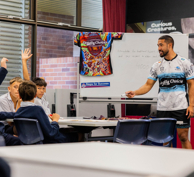 A man in sportswear stands by a whiteboard, engaging with students in a classroom as part of the Deadly Choices Education Programs. Some students raise their hands. A colorful shirt hangs on the board, and educational posters are visible in the background.