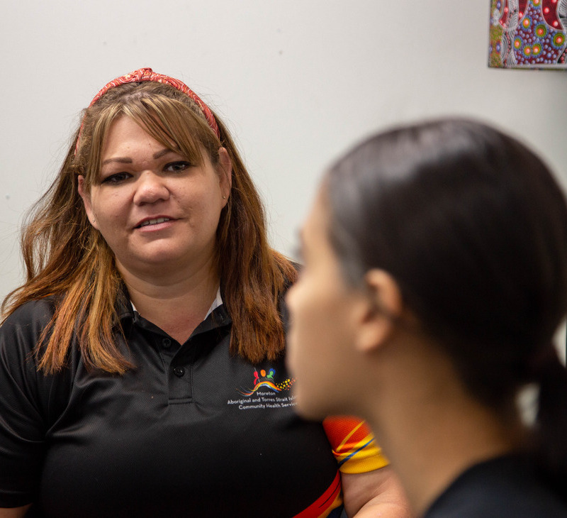 A woman in a black shirt talks to another woman whose face is out of focus. There is colourful artwork on the wall in the background. They are talking about Cervical Cancer Screening.