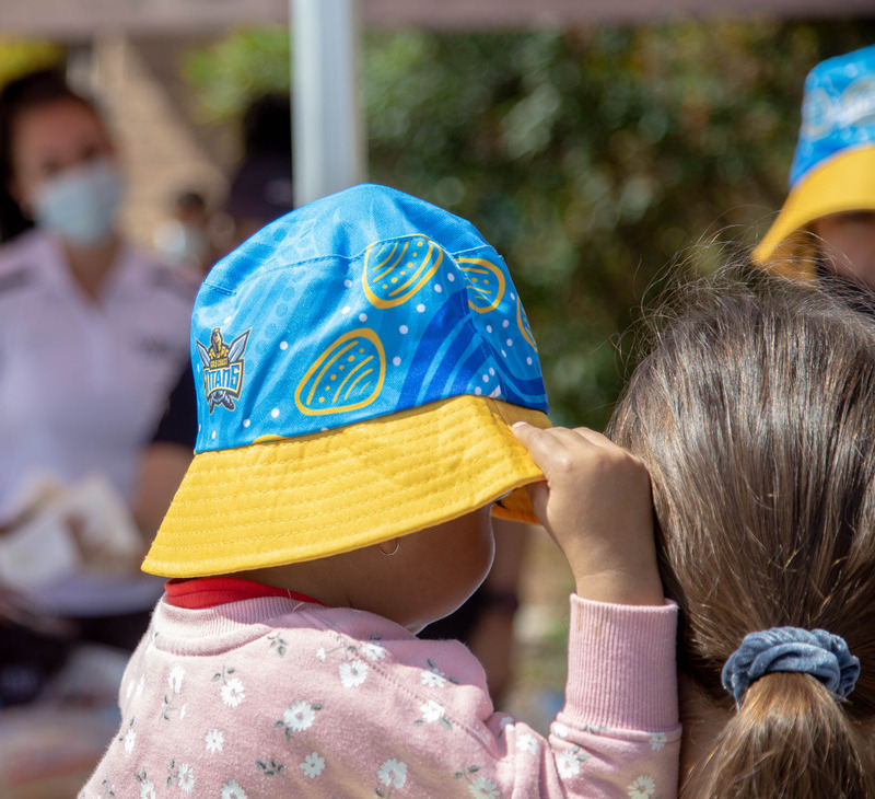A child wearing a blue and yellow hat is held by an adult with brown hair tied in a gray scrunchie. People stand in the blurred background, one wearing a white mask—a common sight during the recent heatwave.