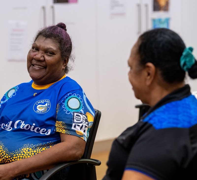 Two women sit indoors, smiling at each other in a warm moment. One, wearing a colorful blue and yellow shirt, sits in the foreground, while the other—perhaps confiding about her experience with menopause—faces away in a blue and black shirt. White cabinets are visible in the background.
