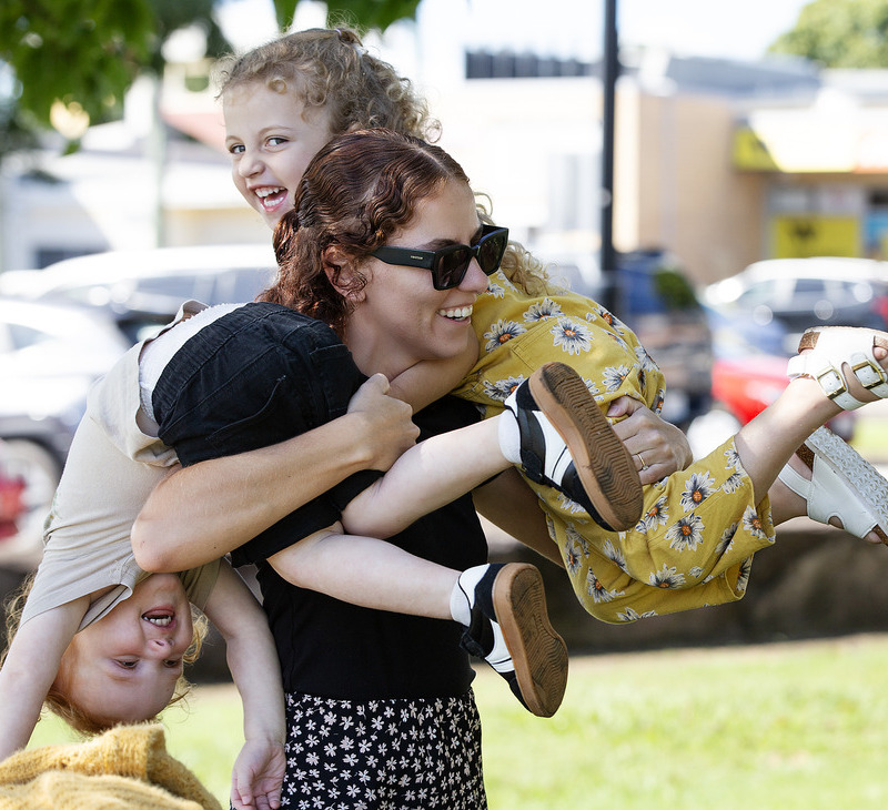 A woman wearing sunglasses carries two laughing children on her shoulders in a playful pose at a sunny park, embodying the joy and connection celebrated by BiOC Mental Health Our Way, with blurred cars and buildings in the background.