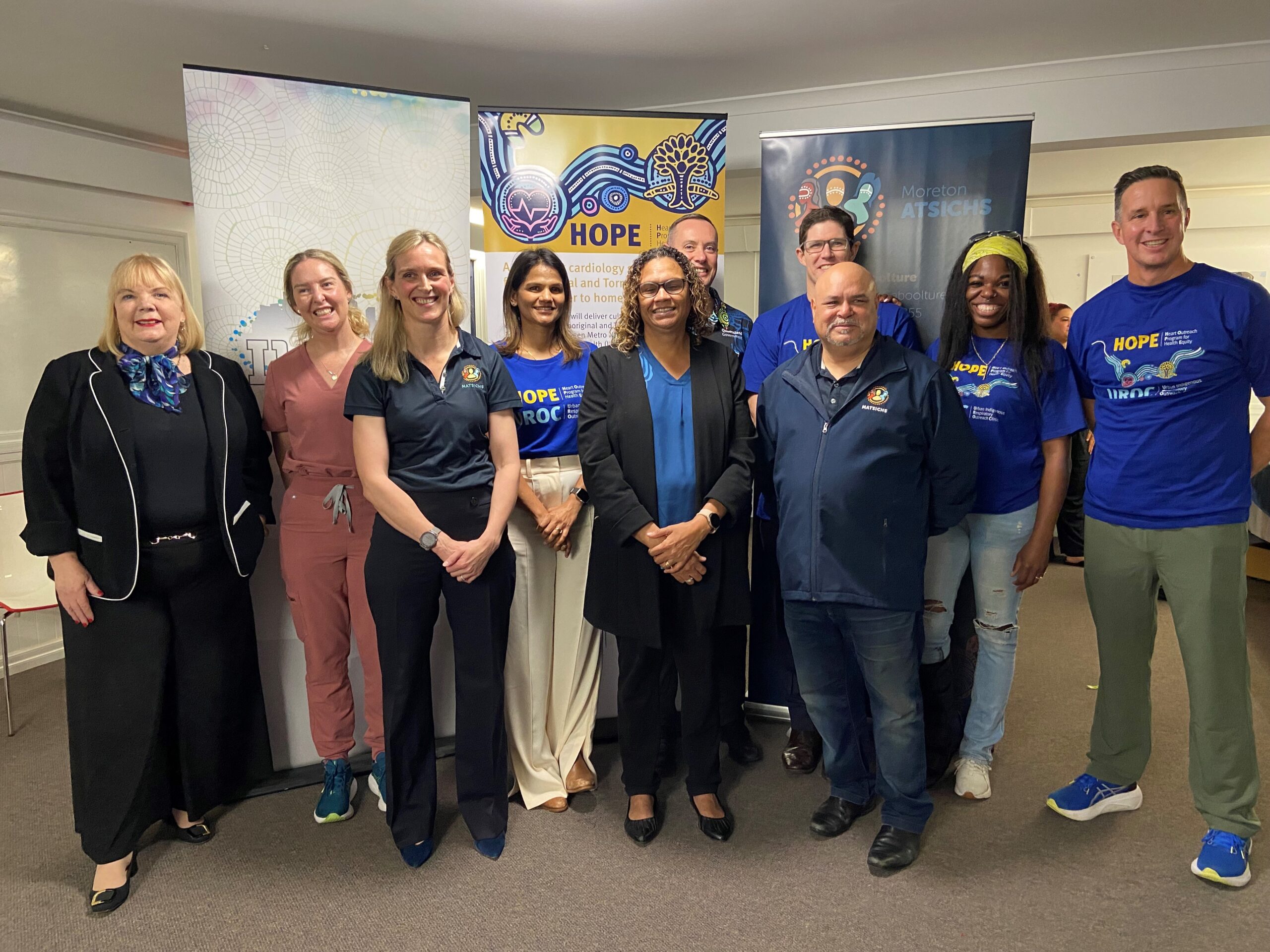 A diverse group of ten people stands and smiles in front of colourful banners at an indoor event, representing HOPE for Mob. Some wear matching blue "HOPE" shirts, while others wear business or casual attire.