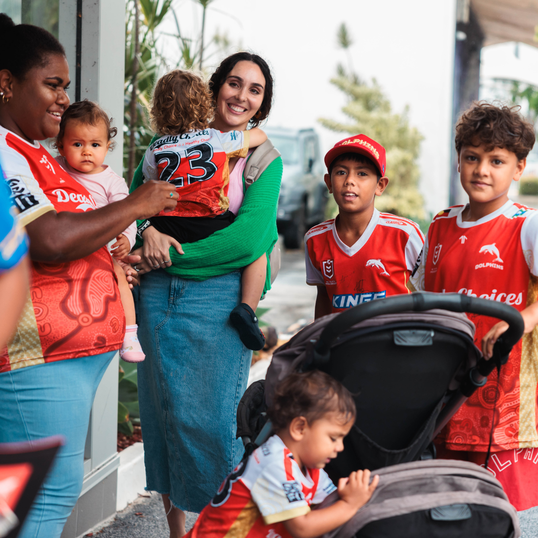 A group of smiling adults and children, some in red sports jerseys, gather outdoors at the Moreton ATSICHS Dolphins Launch Event. One woman holds a toddler, another carries a child, and a young boy sits in a stroller in front.
