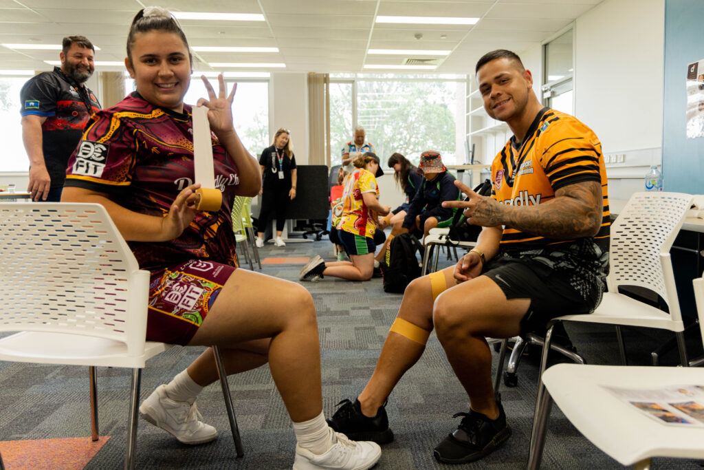 Two people sit facing each other, smiling and holding up peace signs. Wearing colorful sports jerseys, they appear to be members of a Youth Advisory Group. Others in similar attire gather in the background of a bright, modern room.