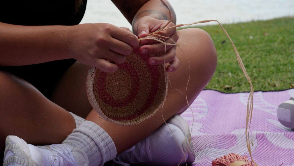 A person from the Healing Hands Women's Group sits cross-legged on a mat outdoors, weaving a small round basket with natural and pink fibers. Their hands hold the basket and weaving materials, and they wear white socks and sneakers on the grass.