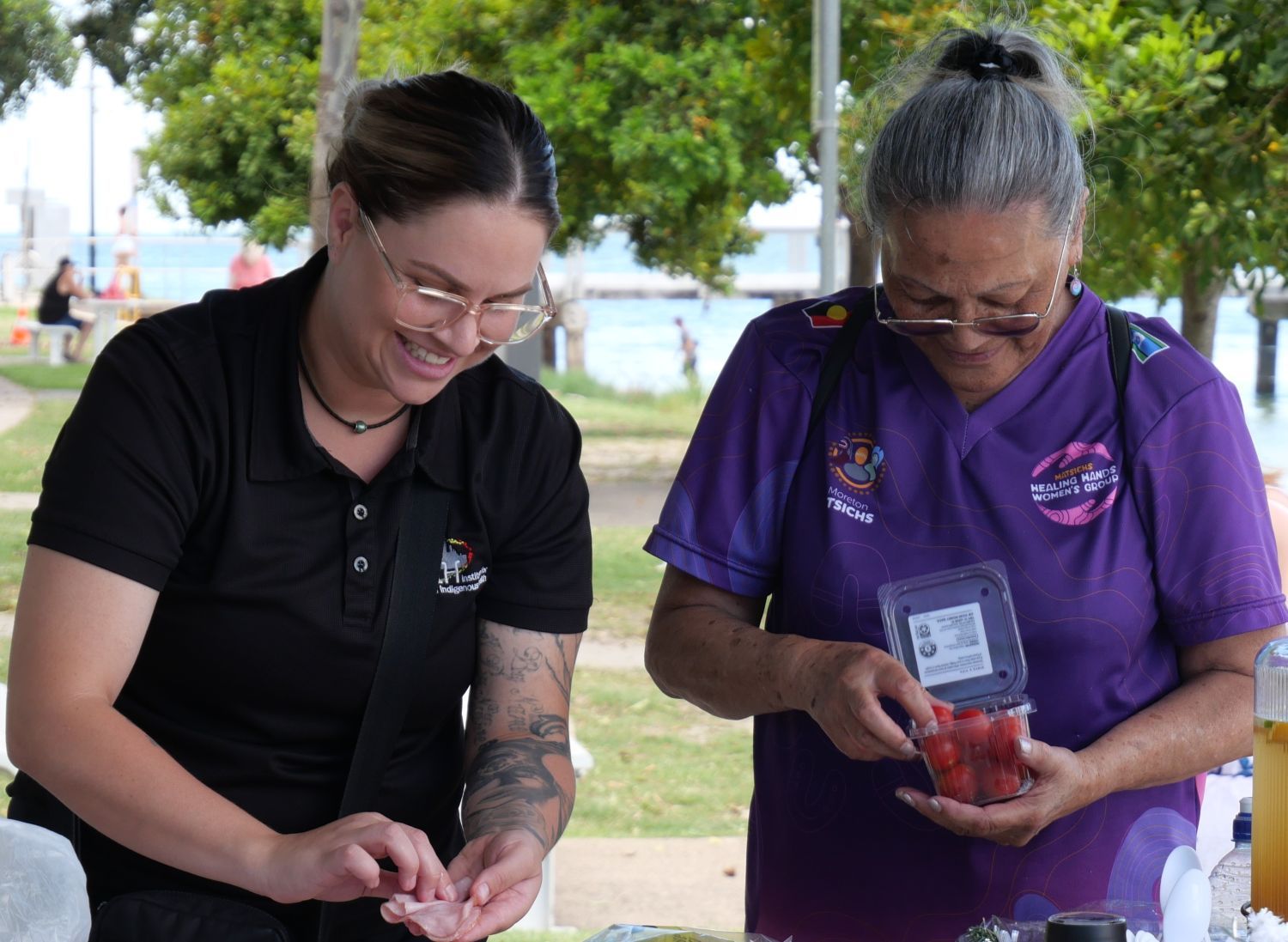 Two women from the Women's Group Healing Hands stand at an outdoor table preparing food. One, wearing glasses and a black Institute for Urban Indigenous Health (IUIH) shirt, is smiling, while the other, in a purple shirt - Healing Hands Group, holds a container of tomatos amid park scenery.