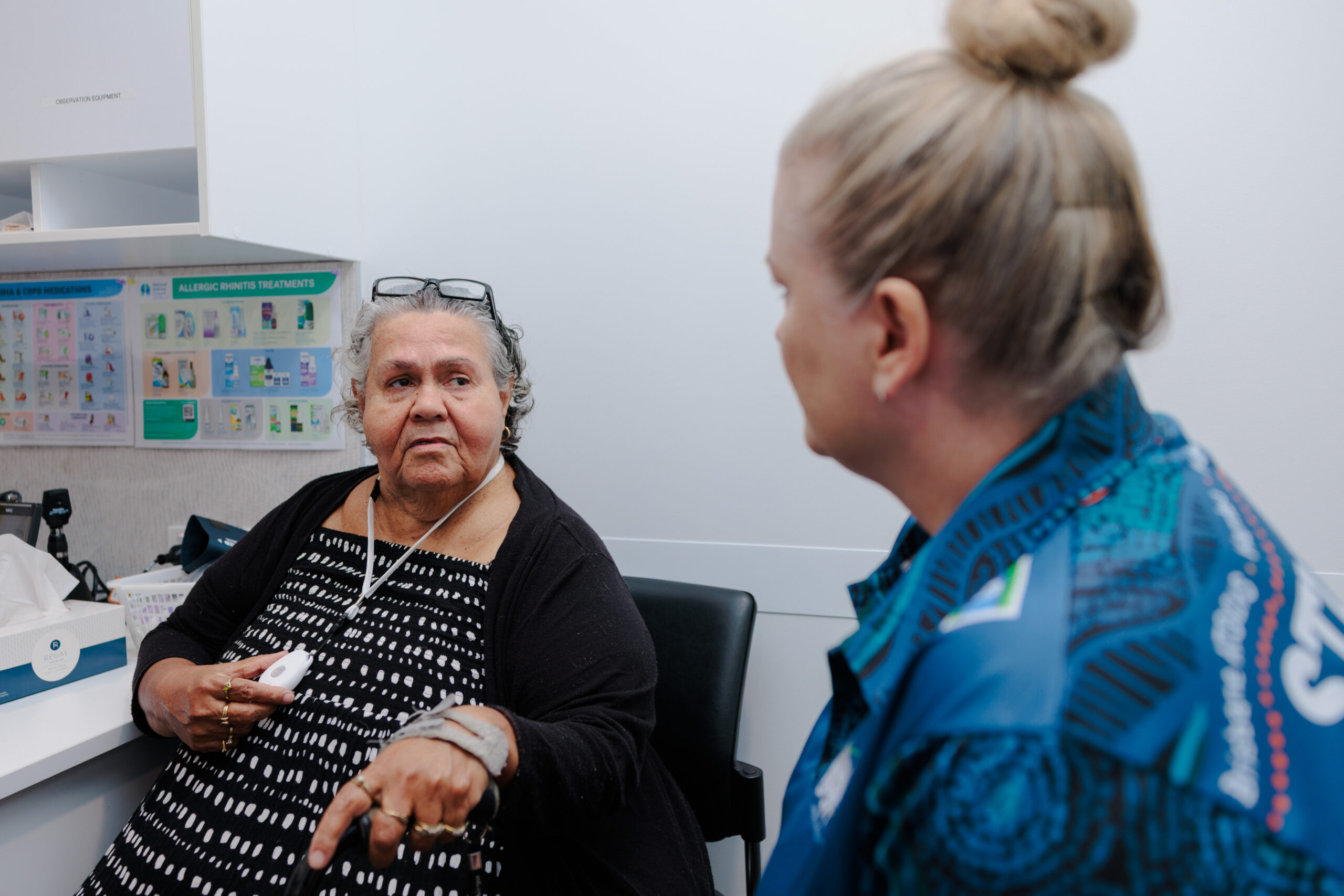 An older woman in a black cardigan sits in a medical office, holding a tissue, and speaks with a healthcare worker in a blue uniform who is listening attentively. Medical posters are visible on the wall behind them.