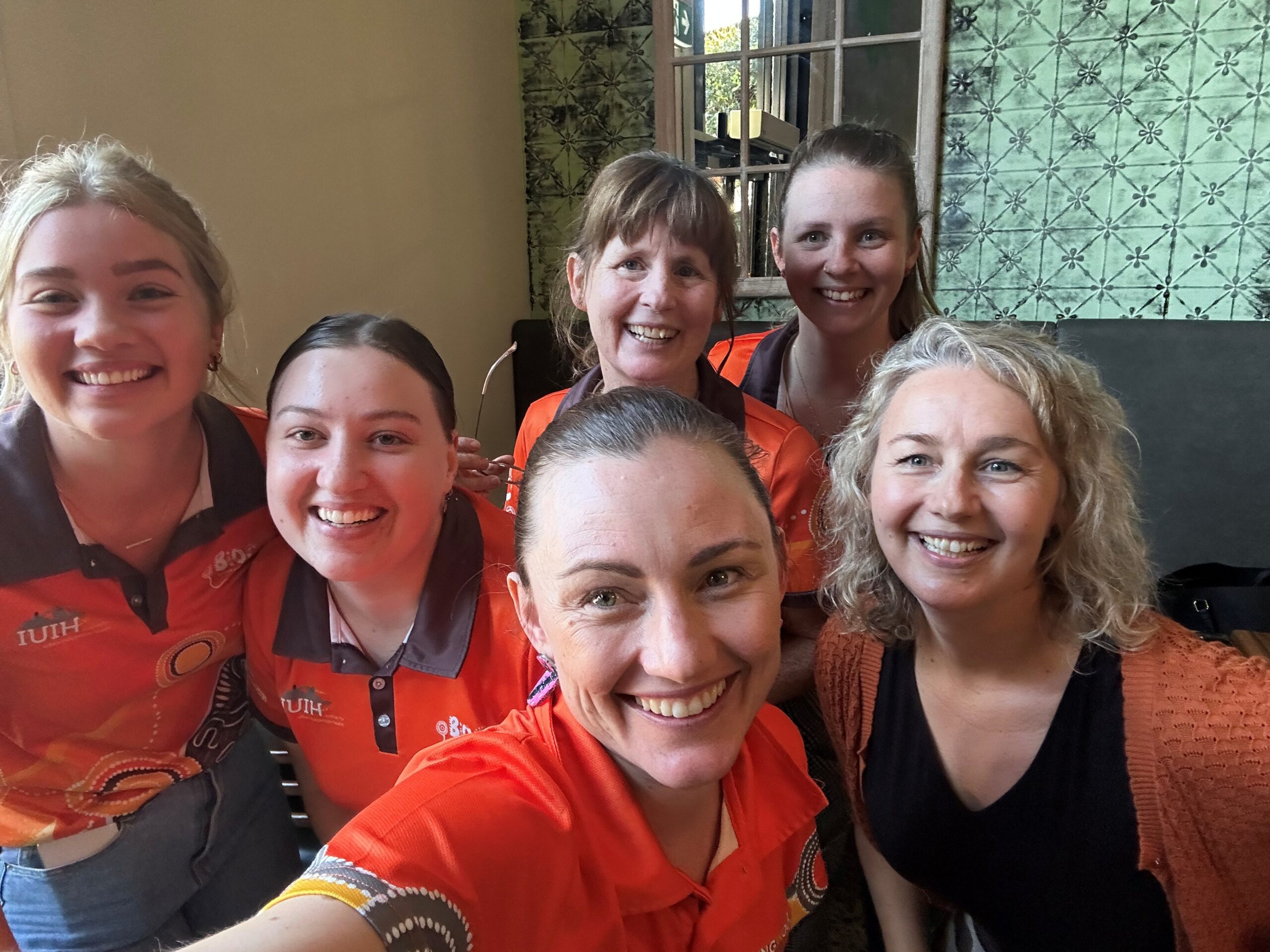 Six midwives smiling and posing for a group selfie indoors, most wearing orange shirts with Indigenous Australian patterns, one wearing a black top with an orange cardigan. They appear happy and cheerful.