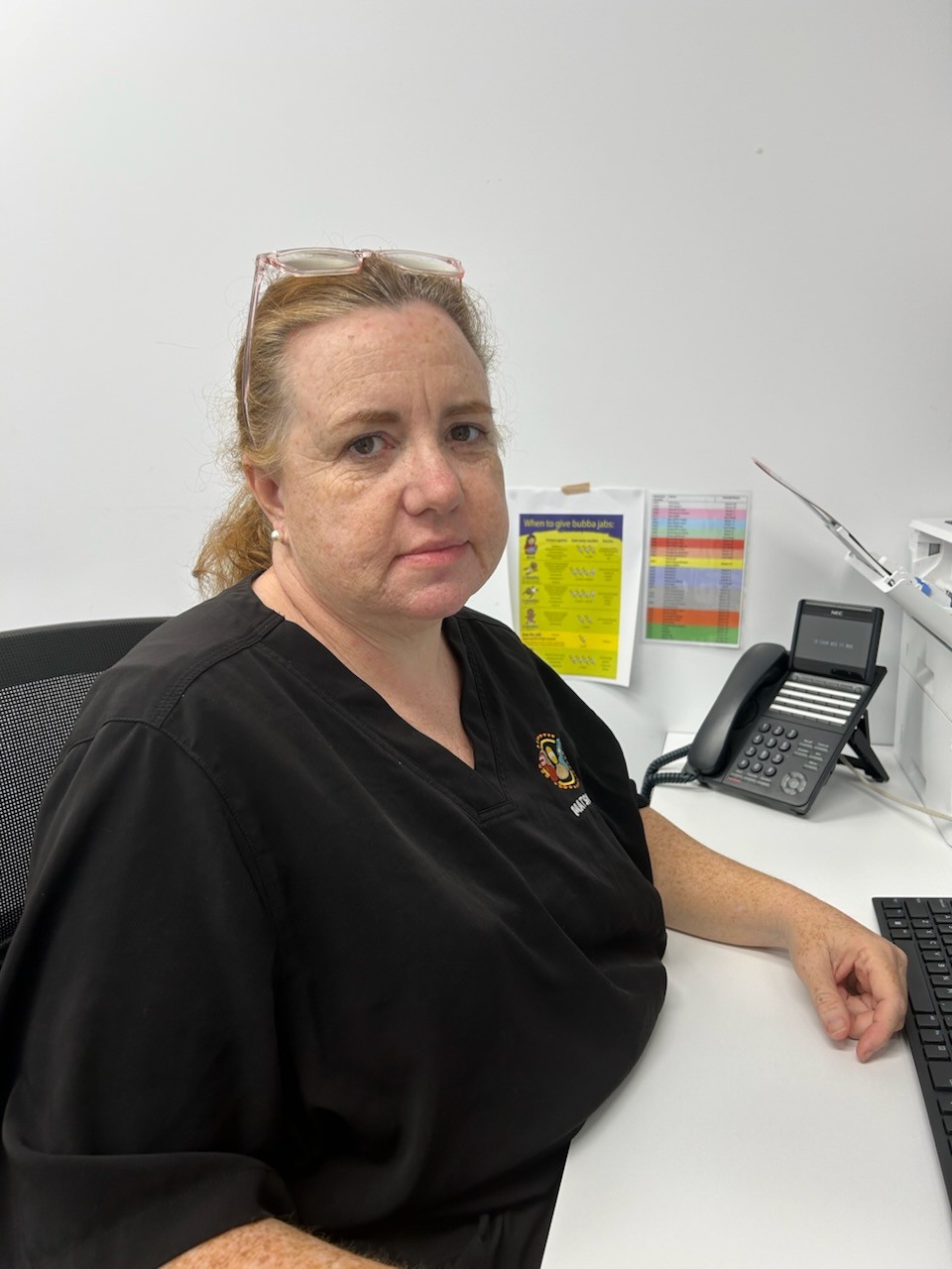 A woman with light brown hair tied back, wearing glasses on her head and a black shirt, sits at a desk with a keyboard, office phone, and colorful charts on the wall behind her, celebrating our nurses and their dedication.
