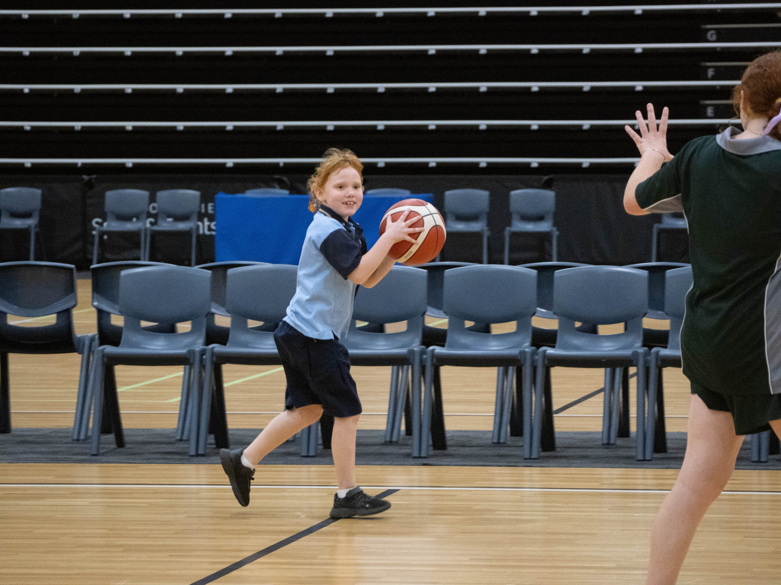 A young child in sportswear, basketball in hand, sprints across an indoor court facing another player.