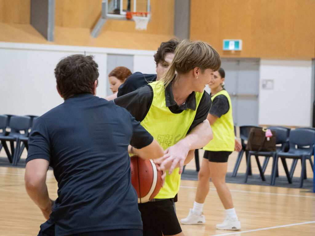 A group of young adults play basketball indoors. One player in a yellow vest dribbles the ball while being closely guarded, as others and empty chairs are seen in the background.