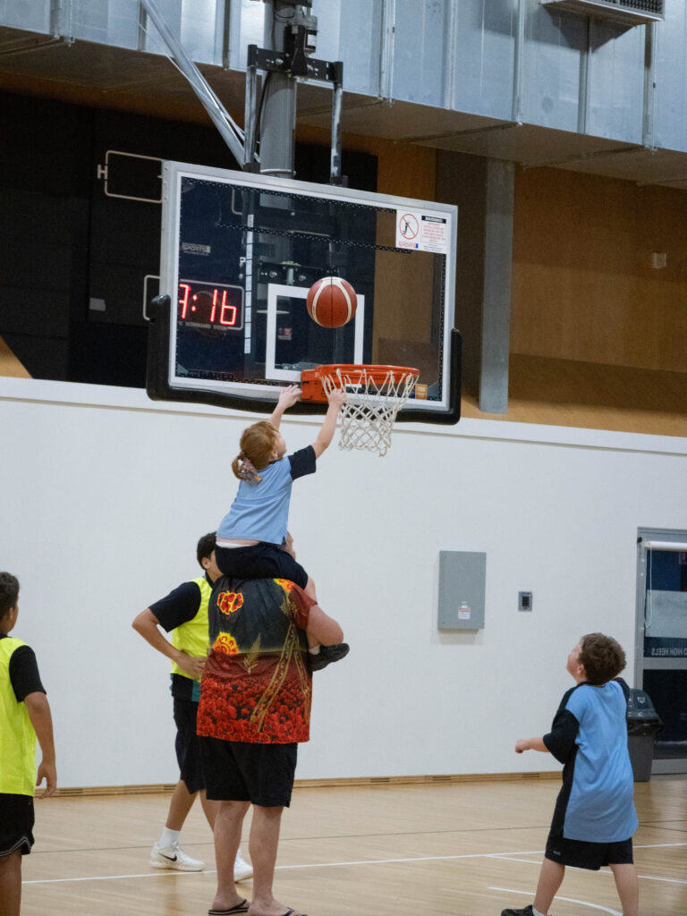 A parent lifts a child towards the basketball hoop as the child attempts a layup.