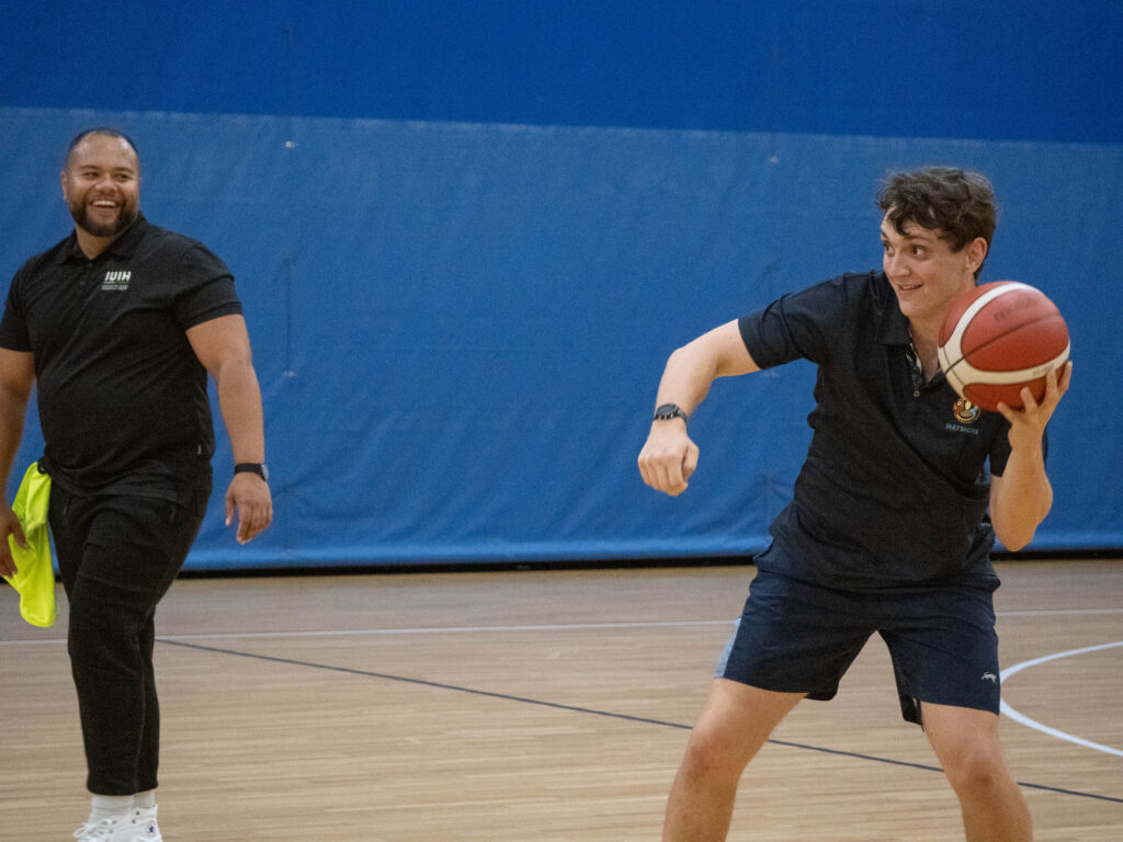Two Moreton ATSICHS staff are on an indoor basketball court. One smiles, watching as the other prepares to throw a basketball.