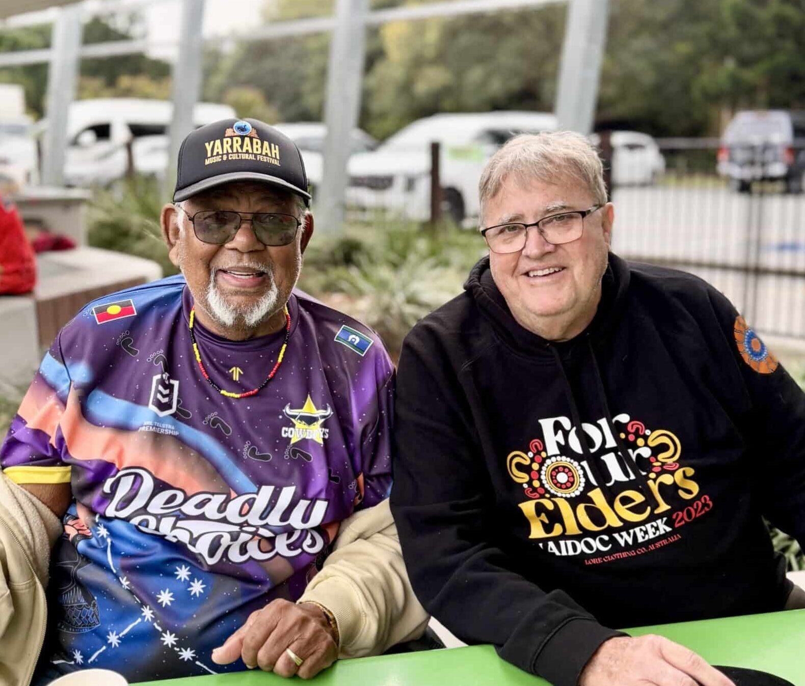 Two men sit at a green table outdoors, smiling at the camera. Their shirts—“Deadly Choices” and “For Our Elders NAIDOC Week 2023”—highlight their support for community-controlled healthcare.