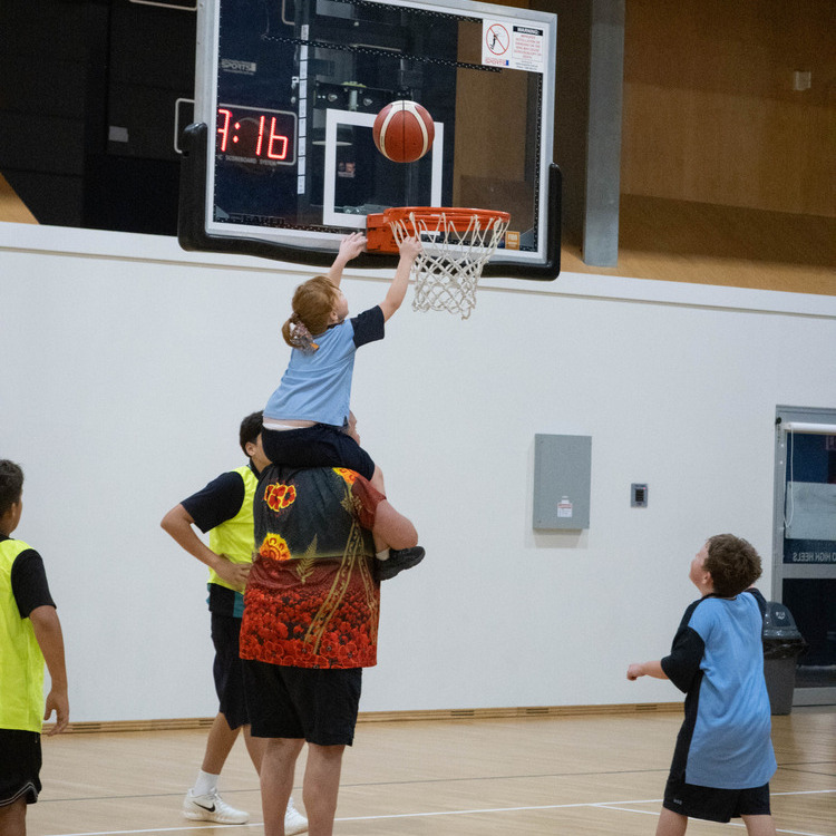 A man holds a young girl on his shoulders as she reaches up to place a basketball into the hoop, chasing her basketball dreams.